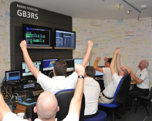 Six men facing a bank of computer screens punch the air in jubilant celebration at GB3RS, the RSGBRSGB National Radio Centre amateur radio station, 