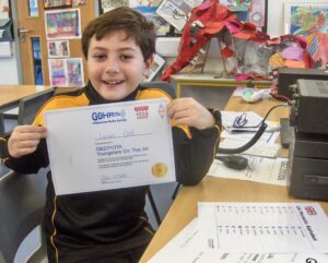 Boy holding up GB23YOTA certificate, sat in front of amateur radio equipment